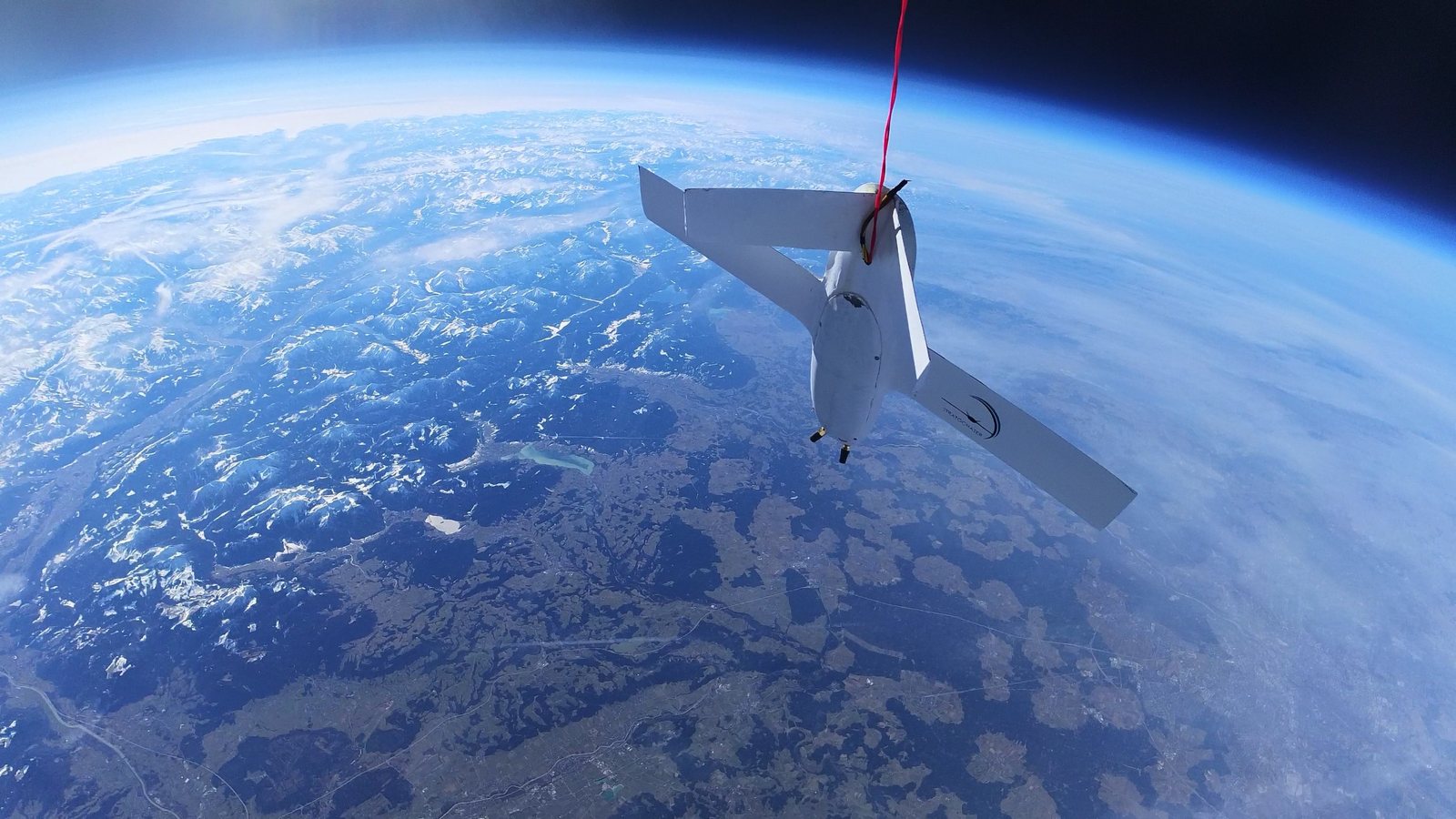 A StratoChaser platform with the StratoChaser logo on its wing, high above southern Germany, with the snow-covered Alps below and the curve of the Earth and the dark sky of the stratosphere on the horizon